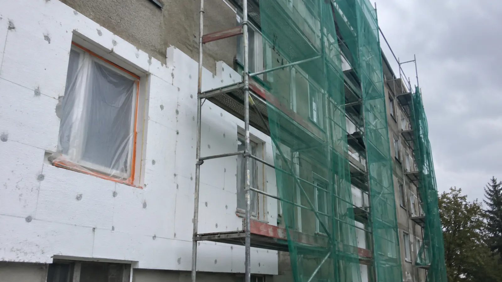 Mehrfamilienhaus bei der Fassadendämmung: halb verlegte weiße Dämmplatten, Gerüst mit grünem Netz, abgeklebtes Fenster
